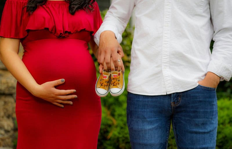 A couple holding baby shoes together. The woman is in a red dress holding her pregnant belly.